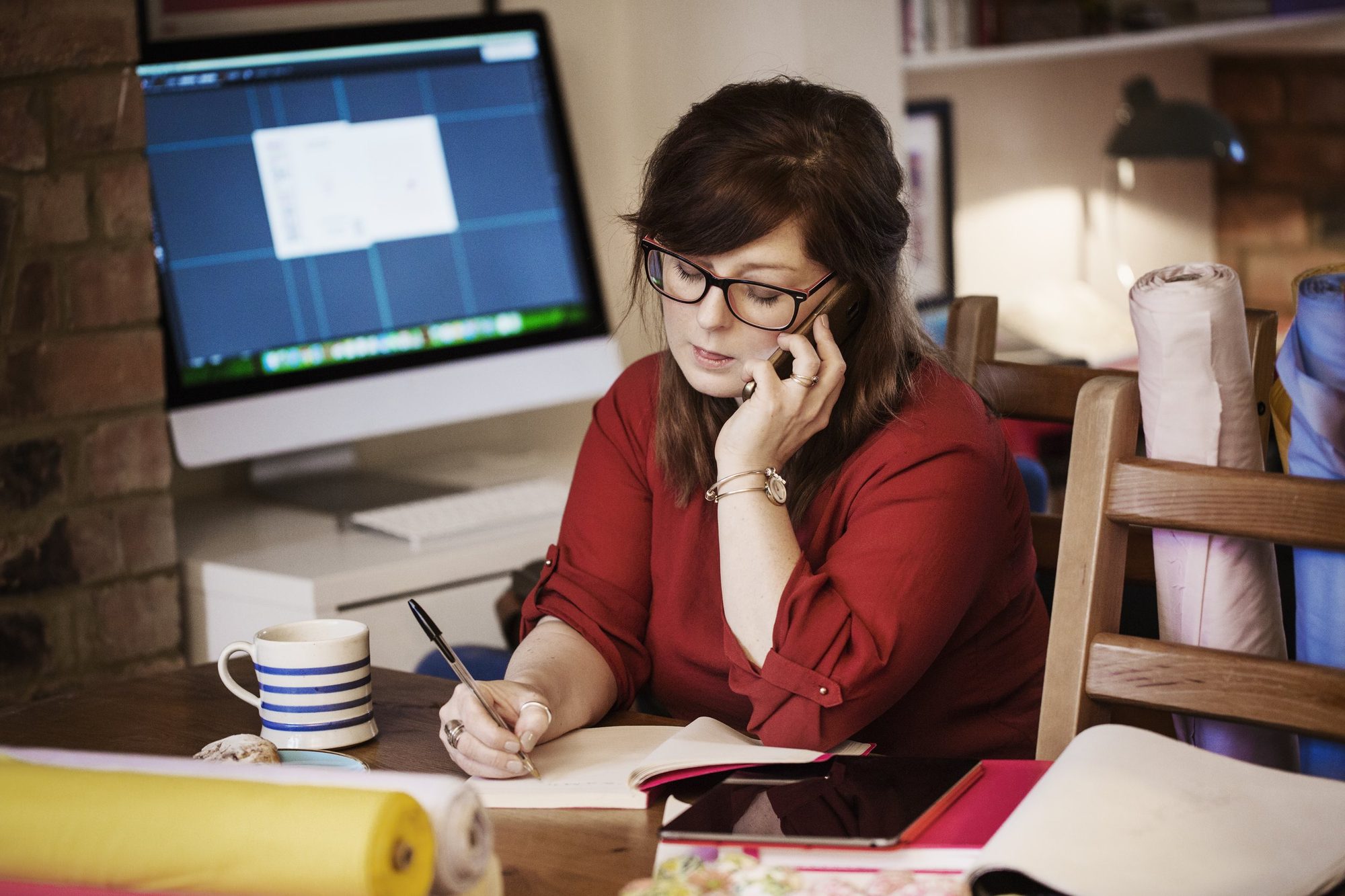 A woman with glasses, brown hair, and wearing a red shirt is sitting at a wooden table talking on a phone, taking notes with a pen, with a laptop, tablet, a coffee mug, and papers in front of her in a cozy home office setting.