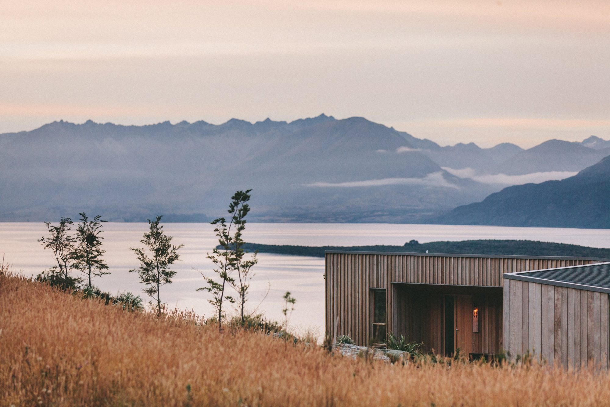 Modern wooden house on a hillside with tall grass, small trees, a large body of water, and mountain range in the background, during sunset or sunrise.