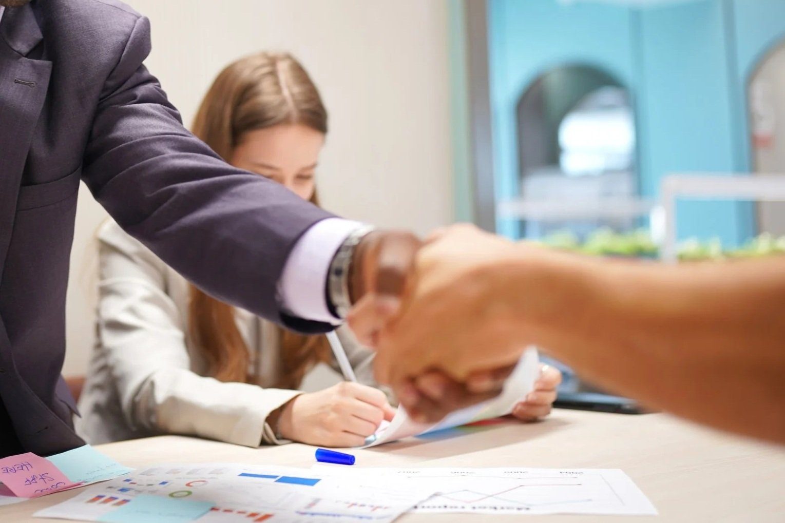 A man in a suit shaking hands with someone, while a woman in the background writes on paper at a table with charts and notes.
