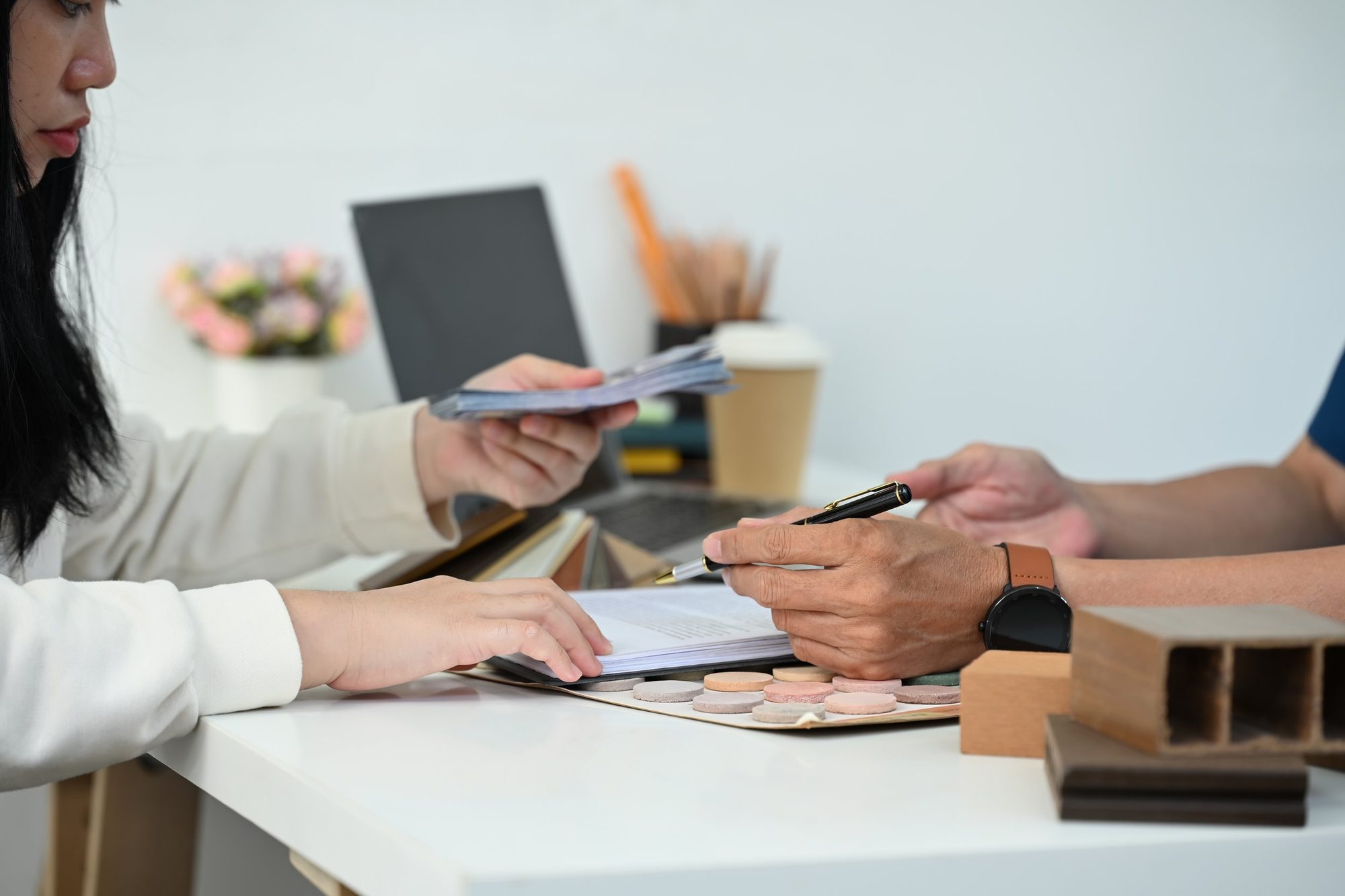 Two people sit at a white table with papers, pens, and color swatches in front of them. One person is handing over a stack of documents, while the other is holding a pen. There are office supplies, a laptop, and a small plant in the background.