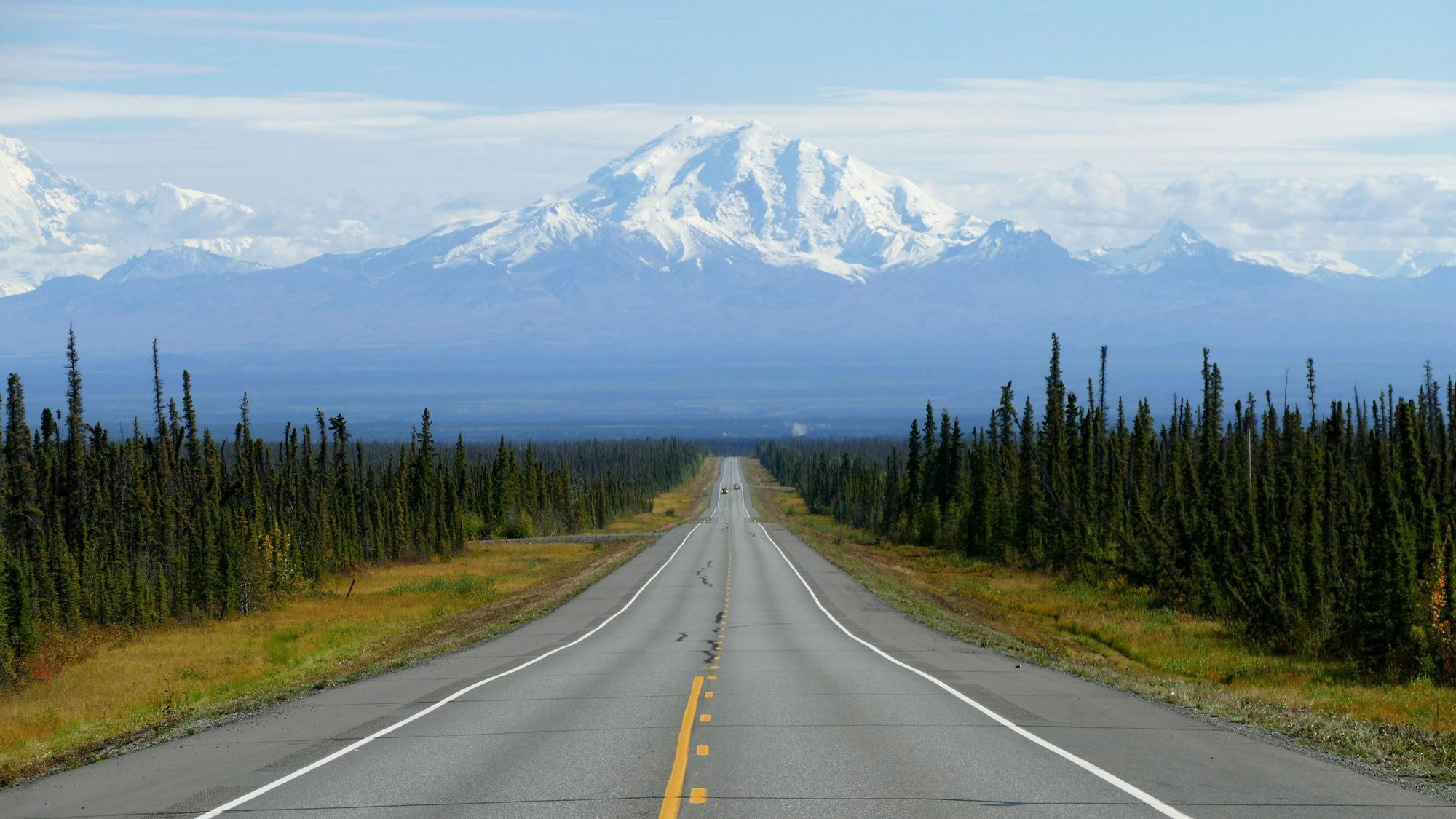 A long, straight road in Alaska with Pioneer Peak in the backround stretching through a forested area with tall, green trees on both sides, leading towards snow-capped mountains in the distance under a partly cloudy sky.