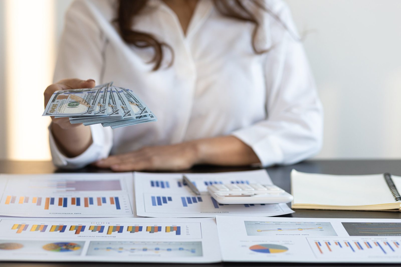 Person holding a fan of hundred-dollar bills on a desk with financial charts, documents, a calculator, and a notebook.